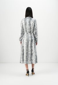 Frau mit glattem, dunklem Haar, die ein langärmeliges, knielanges weißes Kleid mit einem schwarzen Punktmuster und schwarzen High Heels trägt.