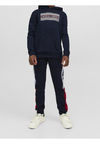 Sudadera azul marino con bolsillo tipo canguro y logo de rayas blancas, combinada con pantalones jogger a juego que presentan acentos rojos y blancos en los laterales.