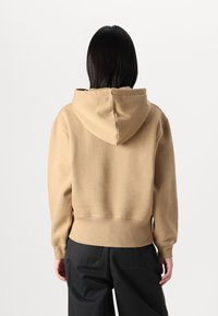 Beige mikina s kapucí a stahovací šnůrkou, vyrobená z měkké tkaniny. Zadní pohled ukazuje jednoduchý design bez viditelných grafik nebo potisků.