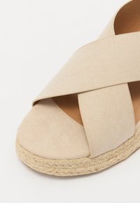 Beige mokasini-korkokenkä, jossa on ristikkäiset hihnat, sileä päällinen ja kudottu juteepohja, pyöreä kärki ja pehmeä pinta.