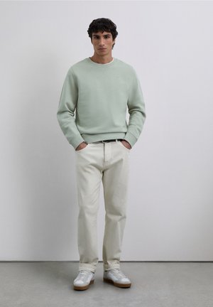 Lichtgroene sweatshirt met geribbelde manchetten en zoom, gecombineerd met gebroken witte broek en witte sneakers met bruine zolen. Effen achtergrond.