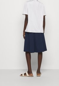 Bílá blůza s krátkým rukávem, plisovaná námořnická sukně a béžové sandály s designem pásku, viděné zezadu na světlém pozadí.