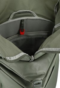 Zaino verde con il compartimento principale aperto, tasca interna grigia visibile, clip di plastica rossa e toppa superiore parzialmente zip.