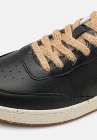 Fekete bőrsneaker lapos jutaszíjjal, perforált orral, varrott varratokkal és foltos textúrájú gumitalppal.