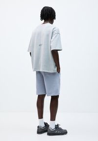 Šedé oversized tričko s jemnou opotřebovanou texturou, spárované se světle modrými šortkami; má černé stehy a šedé sportovní boty.