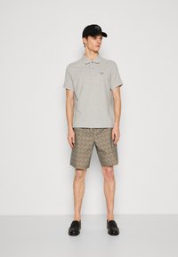 Γκρι πόλο μπλούζα με κοντά μανίκια και διακριτικό λογότυπο, συνδυασμένη με ανοιχτό καφέ σορτς με επαναλαμβανόμενο σχέδιο, και μαύρες λοφάδες.