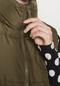 Olivgrön puffer-väst med hög krage och framdragkedja. Utmärkande för sin släta textur och lätta vaddering, bärs över en svart- och vitprickig skjorta.