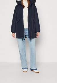Giacca puffa blu navy con cappuccio, dotata di chiusura con zip e tasche laterali, indossata sopra un maglione crema e jeans a gamba larga azzurri.