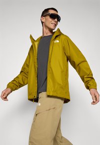 Veste imperméable jaune avec fermeture éclair et poignets ajustables, associée à un pantalon cargo beige avec une poche zippée. Pas de logos de marque visibles.