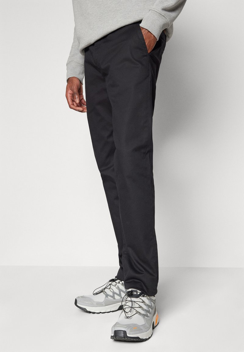 Schwarze Hosen aus einem glatten Material, mit schmaler Passform, seitlichen Taschen und kombiniert mit grauen und orangen Sportschuhen mit strukturierten Details.