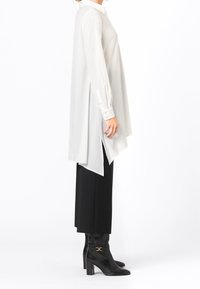 Camicia bianca lunga con colletto a punta, spacchi laterali e maniche lunghe. Abbinata a pantaloni neri aderenti e stivali neri al ginocchio.