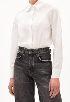 Biała koszula z długim rękawem zapinana na guziki, włożona w czarne wyblakłe dżinsy z wysokim stanem z przednimi kieszeniami i szlufkami na pasek.
