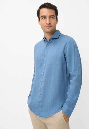 Mann trägt ein hellblaues langärmeliges Hemd mit Knöpfen und beige Hose, steht mit den Händen in den Taschen und schaut leicht nach rechts.