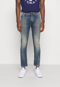 Męskie niebieskie dżinsy z wyblakłym wykończeniem, slim fit i zniszczonymi detalami. W połączeniu z białymi sneakersami i granatowym T-shirtem z grafiką.