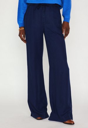 Persoon die marineblauwe wijde broek met elastische tailleband en trekkoord draagt, gecombineerd met bruine puntige schoenen en een blauwe top met lange mouwen.