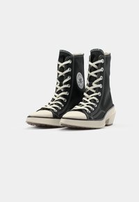 Juodi aukšti sportbačiai su baltais raišteliais, guminiu pirštu ir tekstūruota pode. Ikoniškas logotipo užvalkalas ant šono. Yra kontrastingas siuvinėjimas.