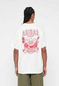 Balta kokvilnas T-krekls ar sarkanu grafiku, kurā attēlots ramen bļoda ar kociņiem, "ADIDAS" teksts un japāņu simboli aizmugurē.