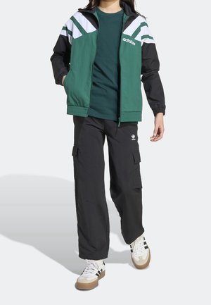 Personne portant une veste Adidas verte et noire, un t-shirt vert foncé, un pantalon cargo noir Adidas et des baskets blanches avec des bandes noires.