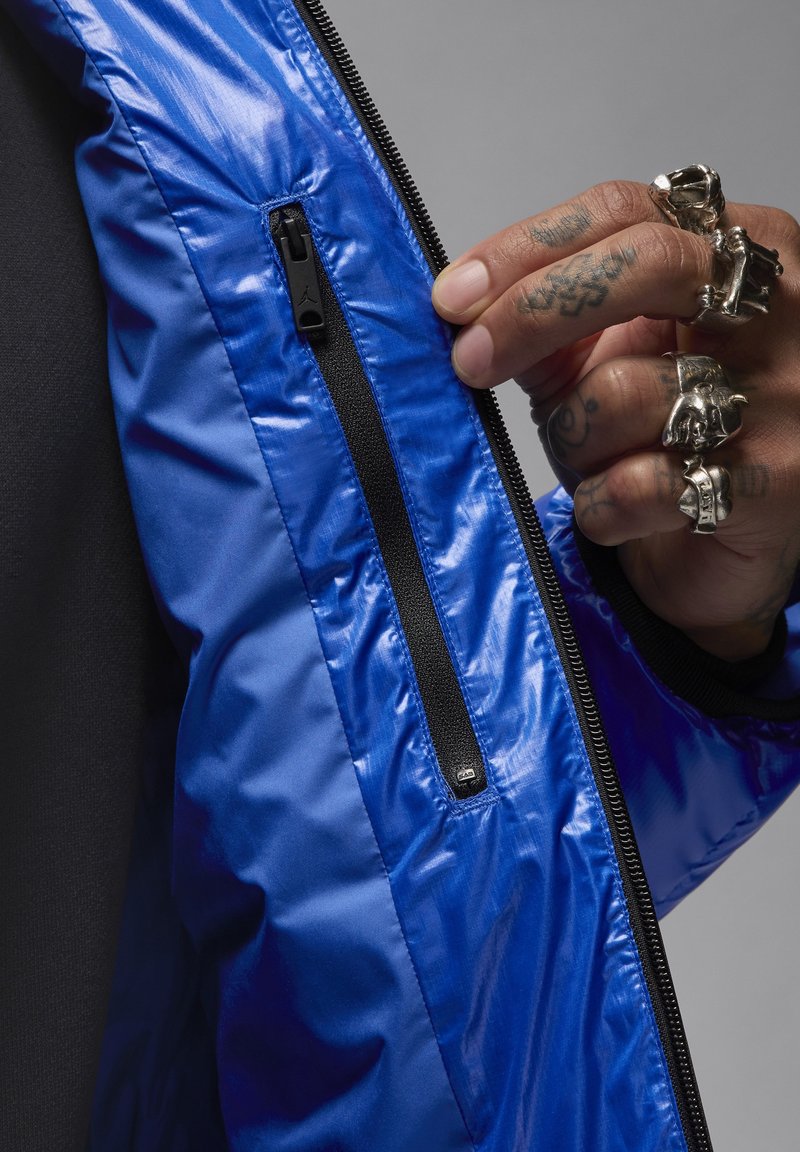 Chaqueta aislante azul con un acabado brillante, que presenta un bolsillo con cremallera y acentos en negro. El material parece suave y ligero.