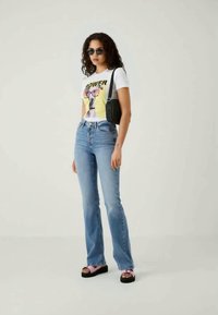 Weißes T-Shirt mit Grafikdruck, hellblaue Schlagjeans, schwarze Handtasche und schwarze Sonnenbrille. Die Sandalen haben einen pinken Riemen und eine schwarze Sohle.