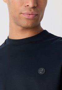 T-shirt bleu marine à manches courtes en tissu doux, avec un col rond et un badge logo circulaire sur la poitrine gauche.