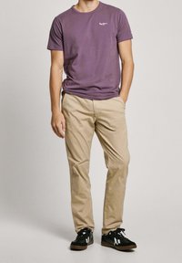 Herre t-shirt med korte ærmer i lilla bomuld med et lille hvidt logo; kombineret med beige bomuldsbukser og sorte sneakers med hvide detaljer.