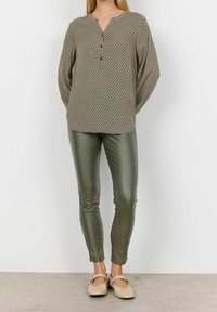 Langärmlige, olivgrün gemusterte Bluse mit einem Spitzkragen und drei Knöpfen, kombiniert mit eng anliegenden, olivgrünen Kunstlederhosen und beigen Ballerinas.