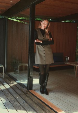 Femme aux longs cheveux portant une robe boutonnée par-dessus un top noir et des bottes, debout les bras croisés dans une pièce en bois près d'une porte vitrée.