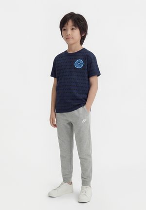Garçon debout les mains dans les poches portant un t-shirt bleu marine à motifs, un pantalon de survêtement gris Nike et des baskets blanches sur fond blanc.