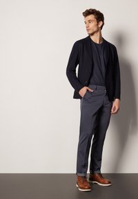 Marinblå blazer över en mörk t-shirt, i kombination med grå byxor och bruna snörstövlar. Slät tygkvalitet med en strukturerad passform och uppvikta byxben.