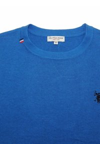 Blå stickad tröja med rundad halsringning, med en liten broderad logotyp och en randig detalj på kragen. Storlek M etikett synlig.