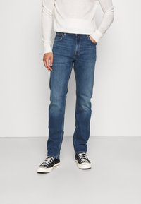 Person wearing blue straight-leg jeans, white long-sleeve shirt, and black low-top sneakers with white laces standing against a plain wall.