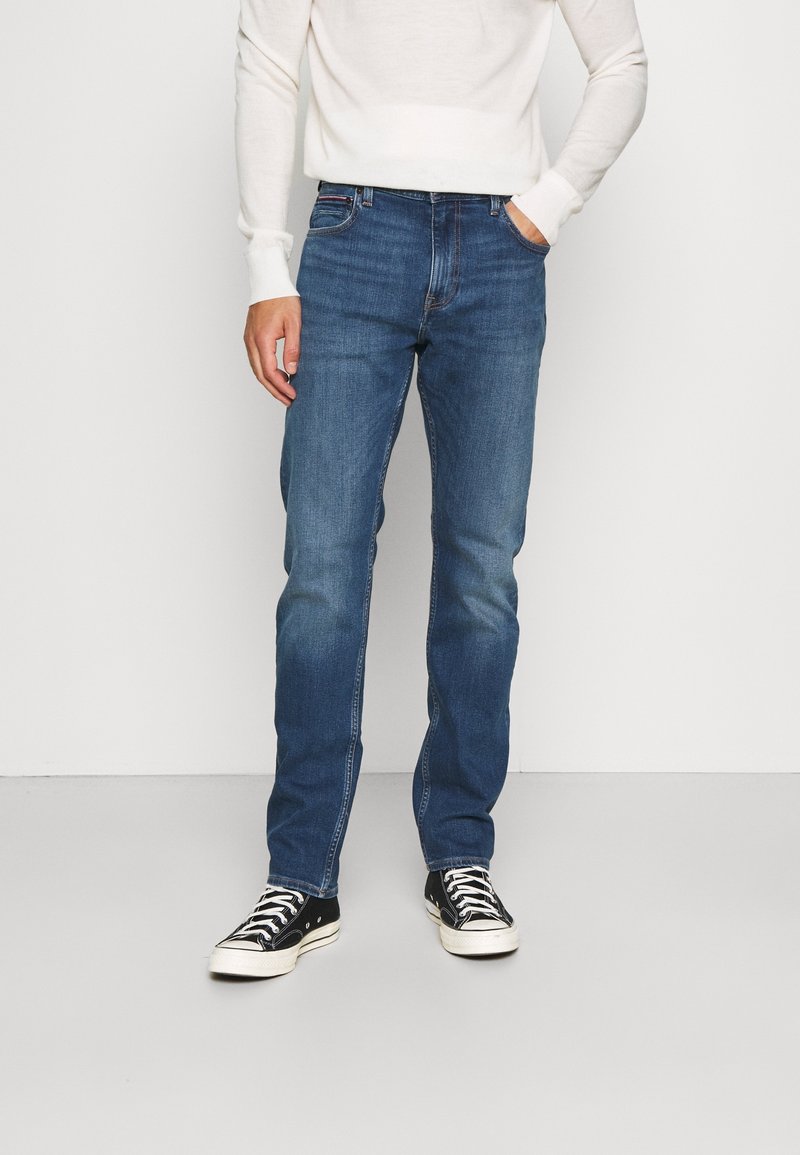 Person wearing blue straight-leg jeans, white long-sleeve shirt, and black low-top sneakers with white laces standing against a plain wall.