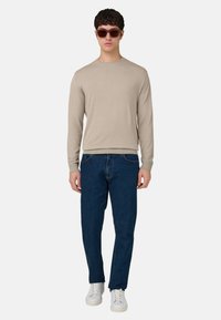 Camicia beige a maniche lunghe, jeans blu scuro e sneakers bianche. Il modello indossa occhiali da sole marroni. Design semplice e vestibilità comoda.