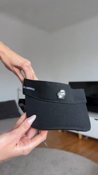 Black visor cap made of fabric, featuring a stitched logo and a small 3D graphic of a character with glasses. Smooth texture, structured design.