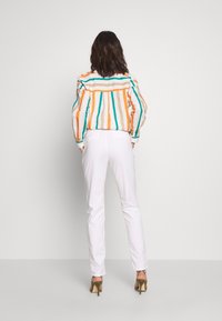 Femme de dos portant une chemise à manches longues rayée multicolore, un pantalon blanc et des sandales à talons hauts vert olive sur un fond uni.