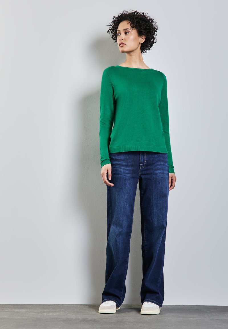 Haut vert à manches longues, encolure ras du cou, associé à un jean droit bleu foncé. Chaussures blanches avec une semelle texturée. Tenue simple et décontractée.