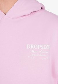 Felpa rosa chiaro con testo bianco che recita "DROPSIZE Street Game Dream Chasers EST. 2020" nella parte alta del petto.