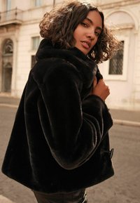 Juodas dirbtinis kailio paltas su aukštu apykakle, priekyje suknelė užsegama mygtukais ir per didelėmis rankovėmis. Lygus paviršius; laisvos, trumpesnės formos.