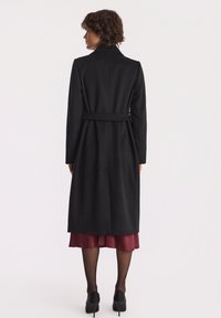 Kobieta widziana z tyłu, ubrana w zapinany na pasek czarny płaszcz na bordową sukienkę, czarne rajstopy i czarne szpilki na białym tle.