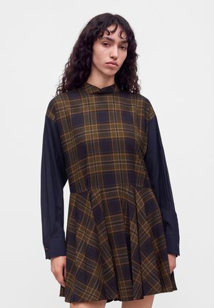 Femme aux longs cheveux bouclés porte une robe à carreaux foncée avec des manches longues unies devant un fond clair uni.