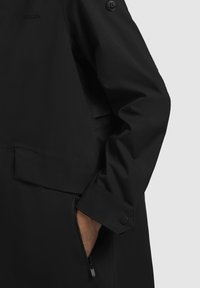 Zwarte waterdichte jas met een soepele textuur, voorzien van een ritszak, knoopsluiting bij de mouwen en een logo-detail op de mouw.