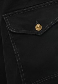 Črna tkaninska žep s beli šivi in zlat gumbom, na katerem je gravura "Versace Jeans Couture" na oblačilu.