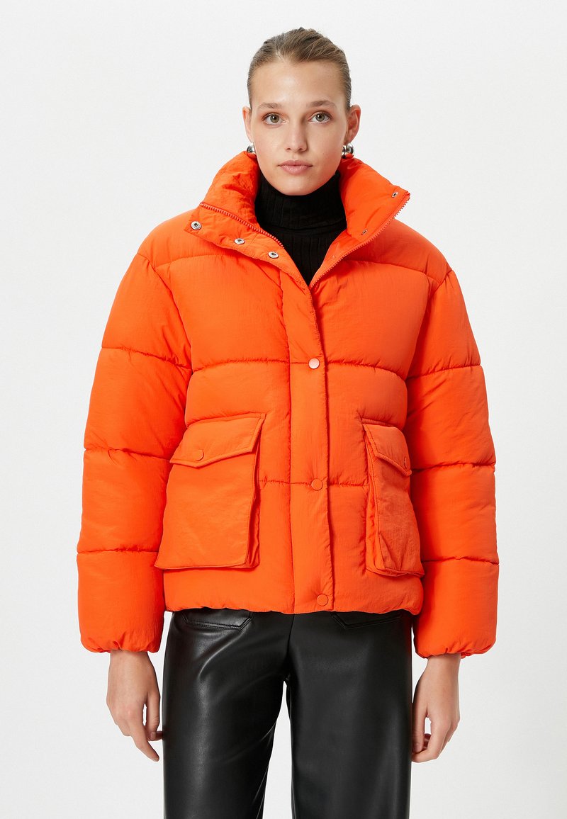 Polstret orange jakke med høj krave, to forlommer og trykknapper. Lavet af et glat stof med en blank finish.