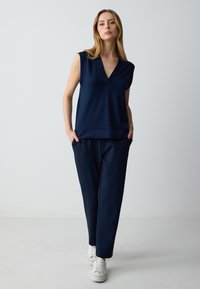 Marineblauw mouwloos topje met een V-hals, gecombineerd met bijpassende marineblauwe tapered pantalons. Gladde stof met zijzakken en witte sneakers.