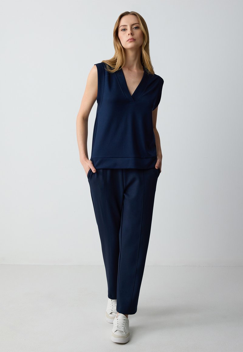 Marineblauw mouwloos topje met een V-hals, gecombineerd met bijpassende marineblauwe tapered pantalons. Gladde stof met zijzakken en witte sneakers.
