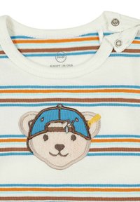 Chemise rayée blanche avec des lignes bleues, oranges et brunes ; présente un visage d'ours brodé portant une casquette bleue avec un détail jaune.