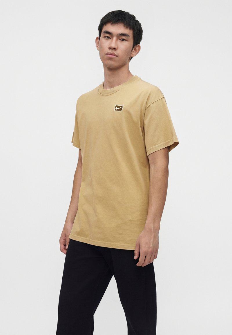 Junger Mann mit kurzen dunklen Haaren, der ein beiges Nike-T-Shirt und schwarze Hosen trägt, steht vor einem schlichten weißen Hintergrund.