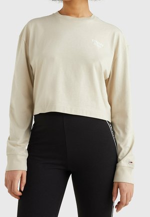 Modèle portant un haut court à manches longues crème Tommy Jeans et un legging noir, sur un fond clair uni.
