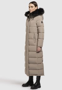 Beige hosszú puffermellény, műszőrme bélelt kapucnival, steppelt mintával, oldalzsebekkel és strapabíró fekete cipzárral. Fekete bokacsizmával viselve.